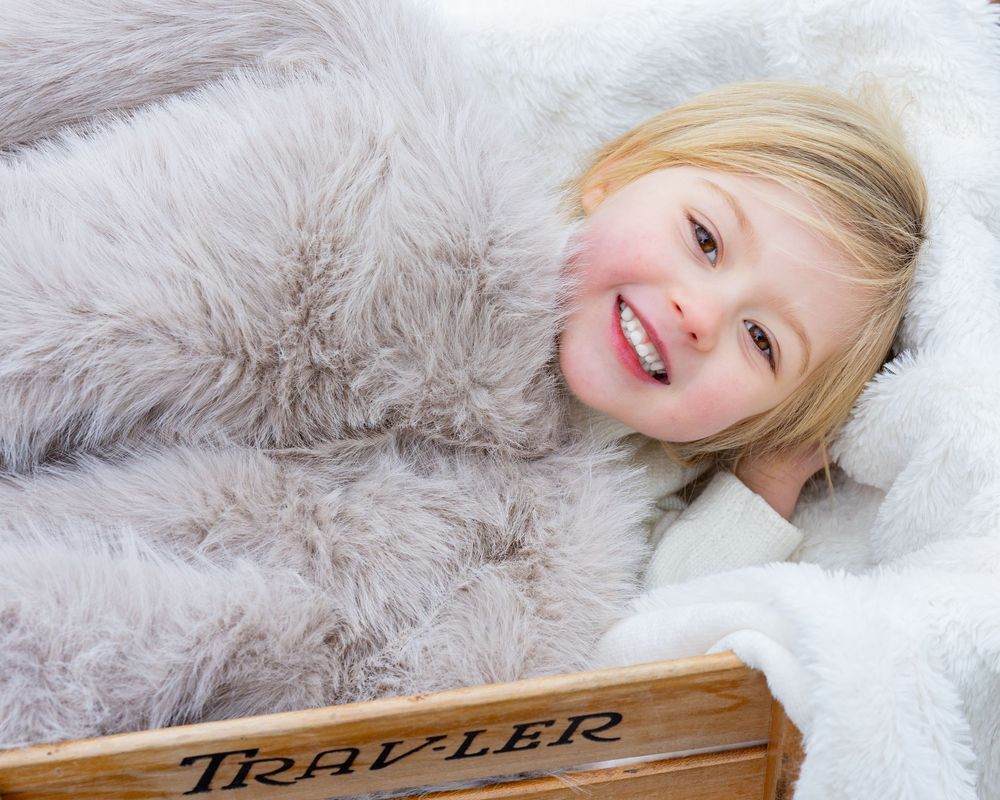 A child with blonde hair lies wrapped in fur bedding in a wagon & smiling brightly during her family photo session in Concord, MA.