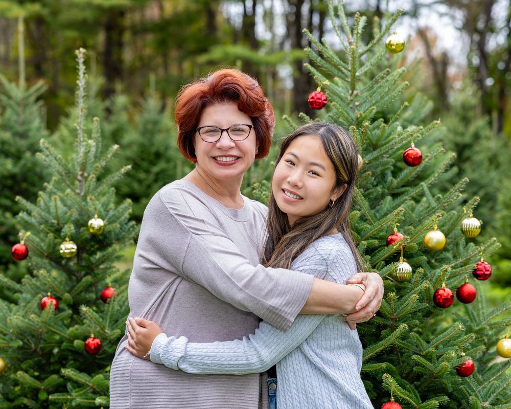 Mother & daughter embrace while standing among decorated Christmas trees outdoors in a festive holiday setting for family photos in Stow, MA