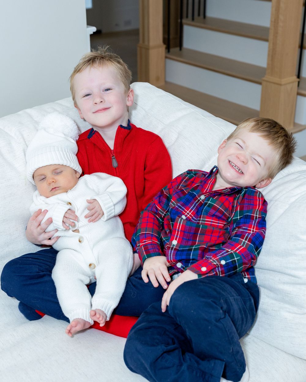 Young siblings children in winter clothing rest together on a white couch during their family's newborn photography session in Sudbury, MA.