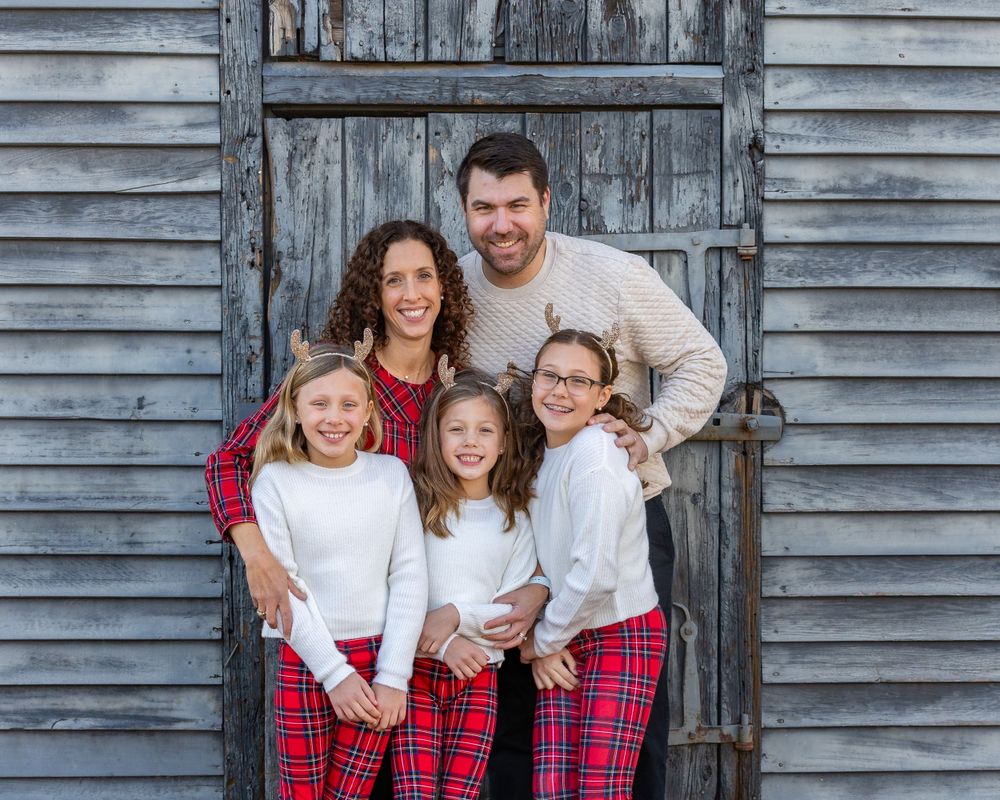 A family poses together in matching holiday outfits in front of a rustic gray wooden barn door at their family photo session in Sudbury, MA.
