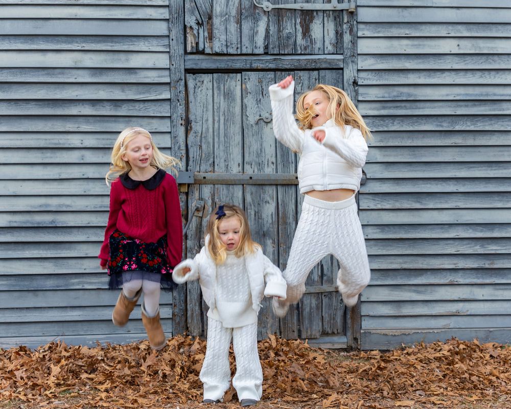 Three sisters in winter outfits dance and jump playfully in front of a rustic gray barn wall at their family photo session in Sudbury, MA.