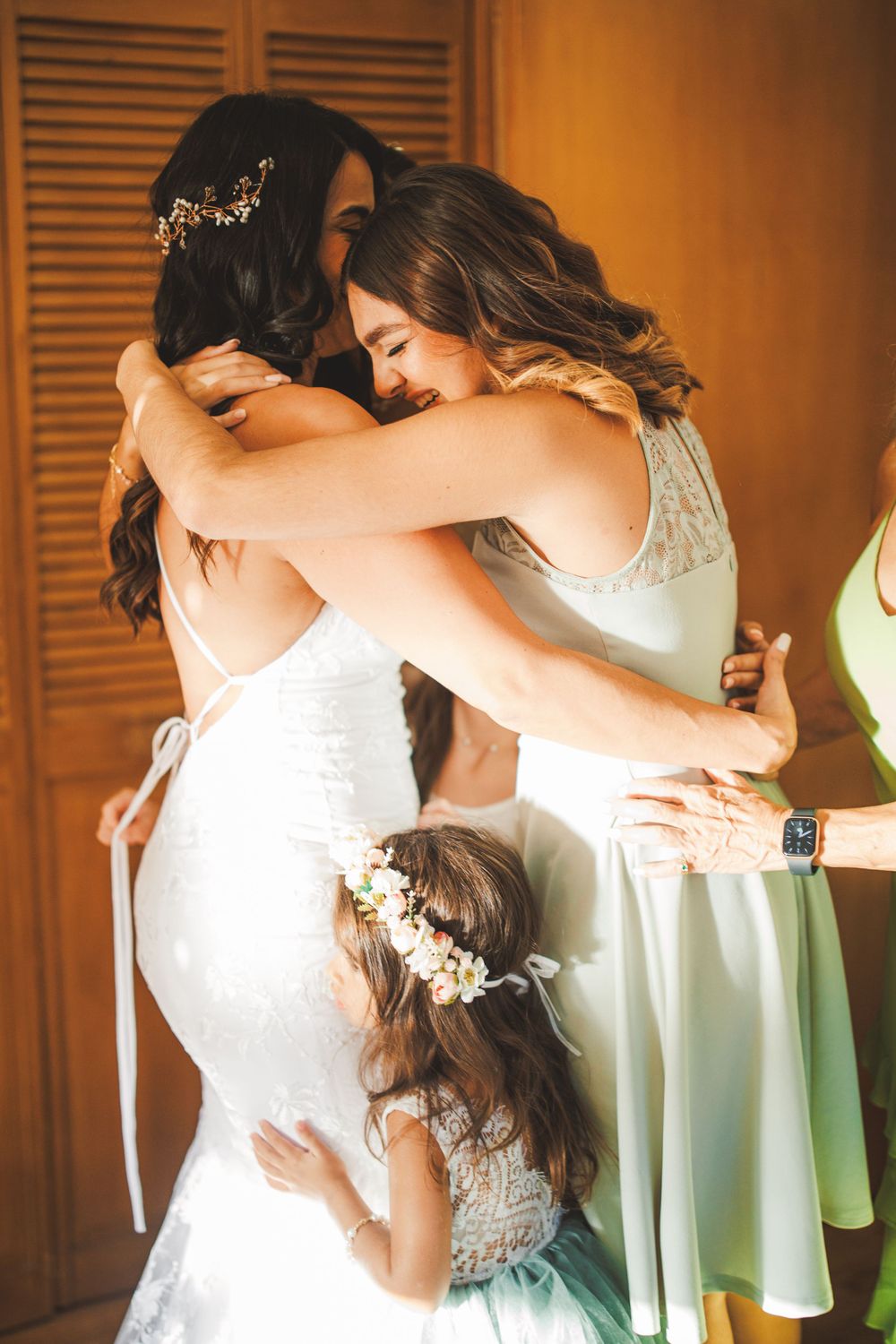 An emotional embrace between women in white dresses with a flower girl joining the moment.