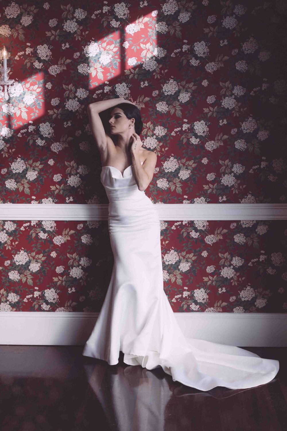 Bride in flowing white gown poses dramatically against floral wallpaper with red lighting accents.