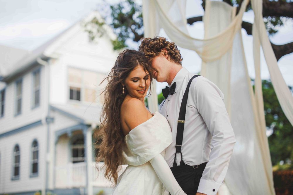 Wedding couple in elegant white dress and suspenders embrace near white building.