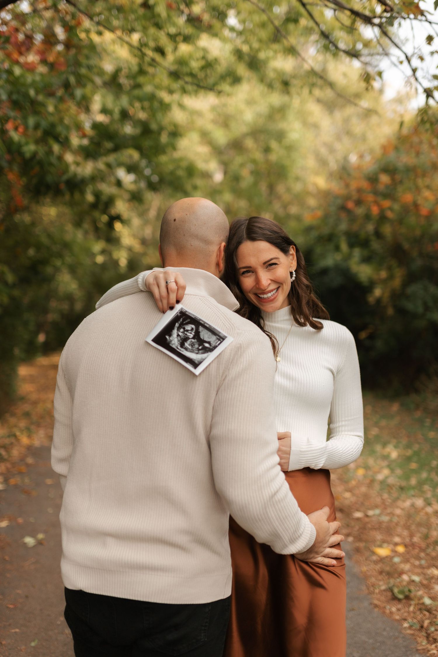 Smiling pregnant woman embracing partner from behind in autumn park while he holds ultrasound photos.