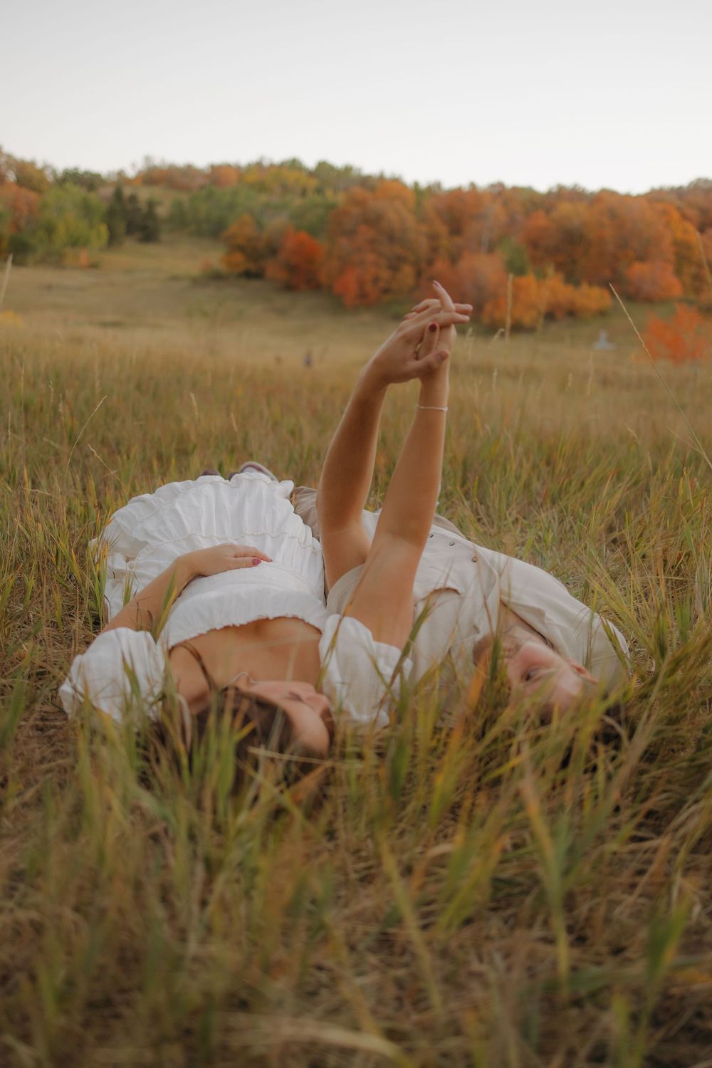 Woman in a white dress lies in a golden autumn meadow with colorful fall foliage in the background.