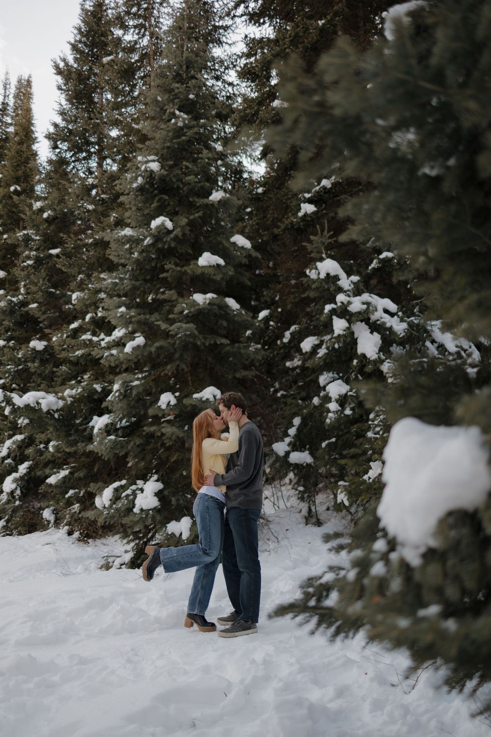 Couple shares a romantic kiss in a snowy winter forest surrounded by snow-covered pine trees.