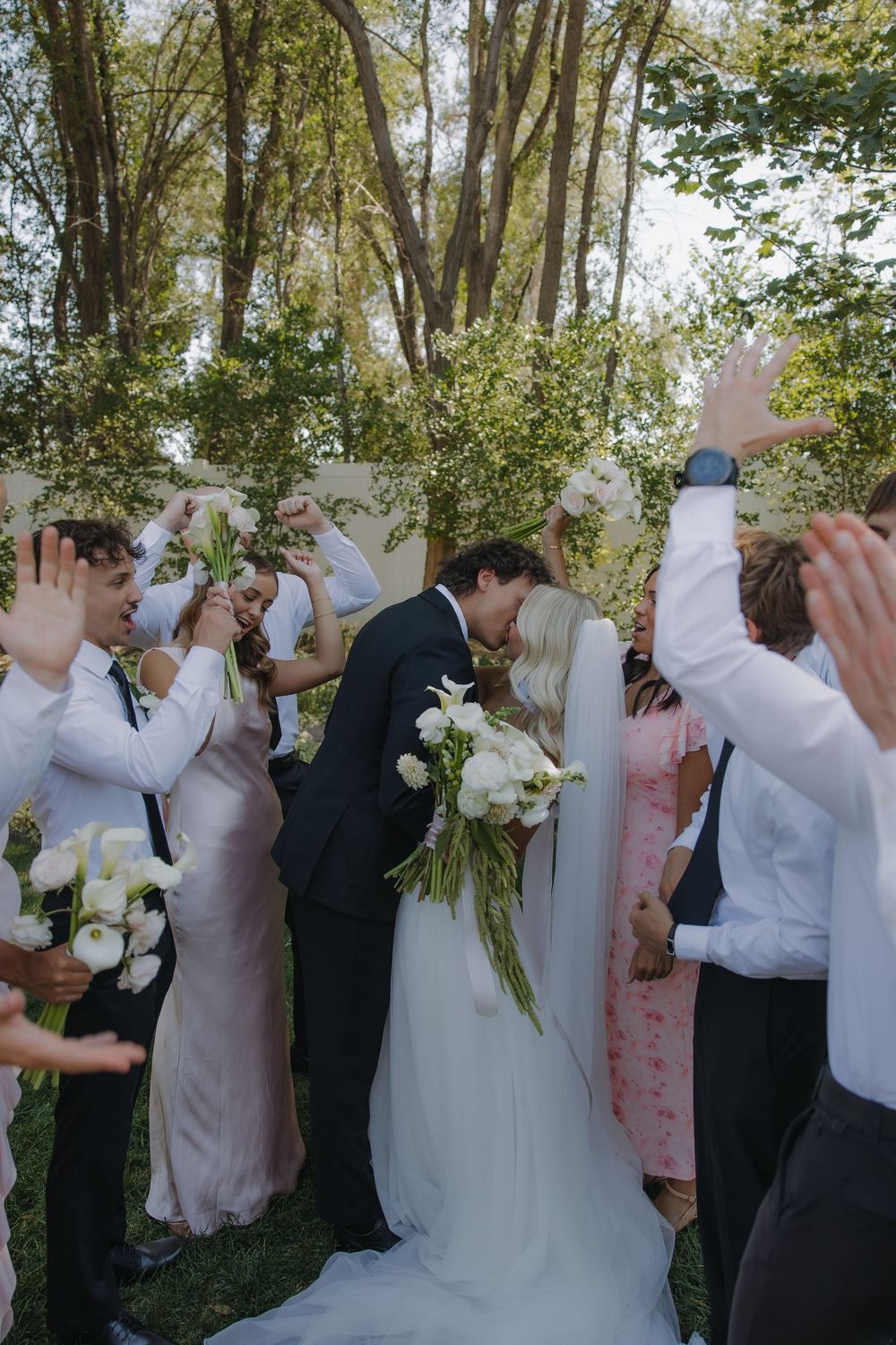 Bride and groom kiss at the altar as wedding party cheers with raised hands in an outdoor ceremony.