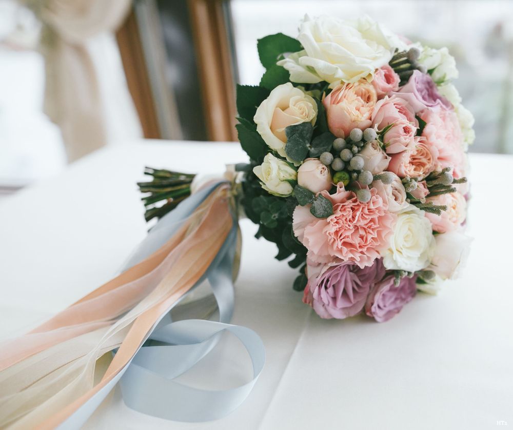 A romantic wedding bouquet with blush pink roses, white carnations, and trailing peach and gray ribbons rests on a table.
