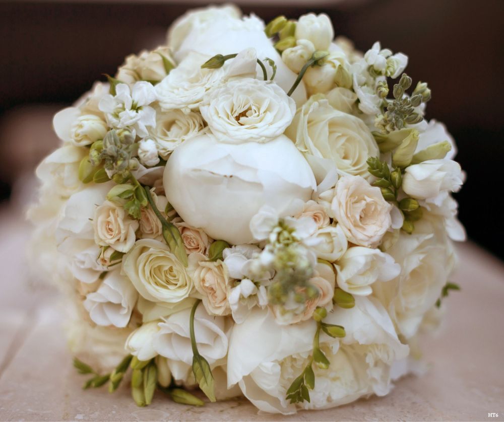 A stunning bridal bouquet featuring cream-colored roses, white peonies, and delicate green foliage in a round arrangement.