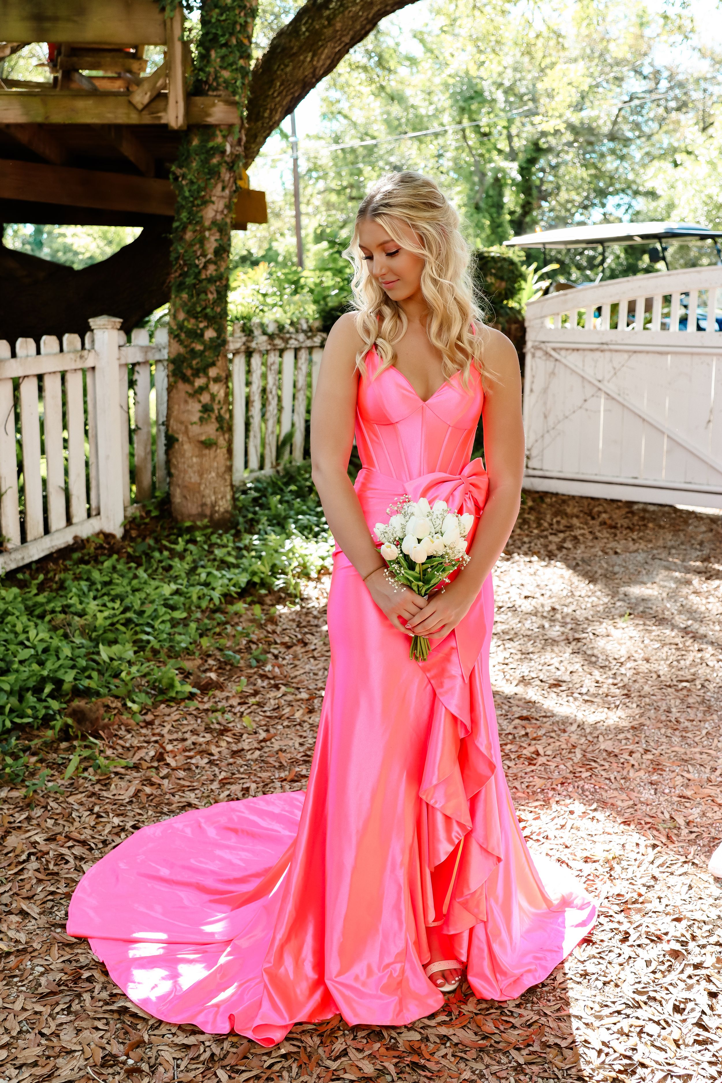 Person in flowing coral pink formal gown holds white bouquet in garden setting.