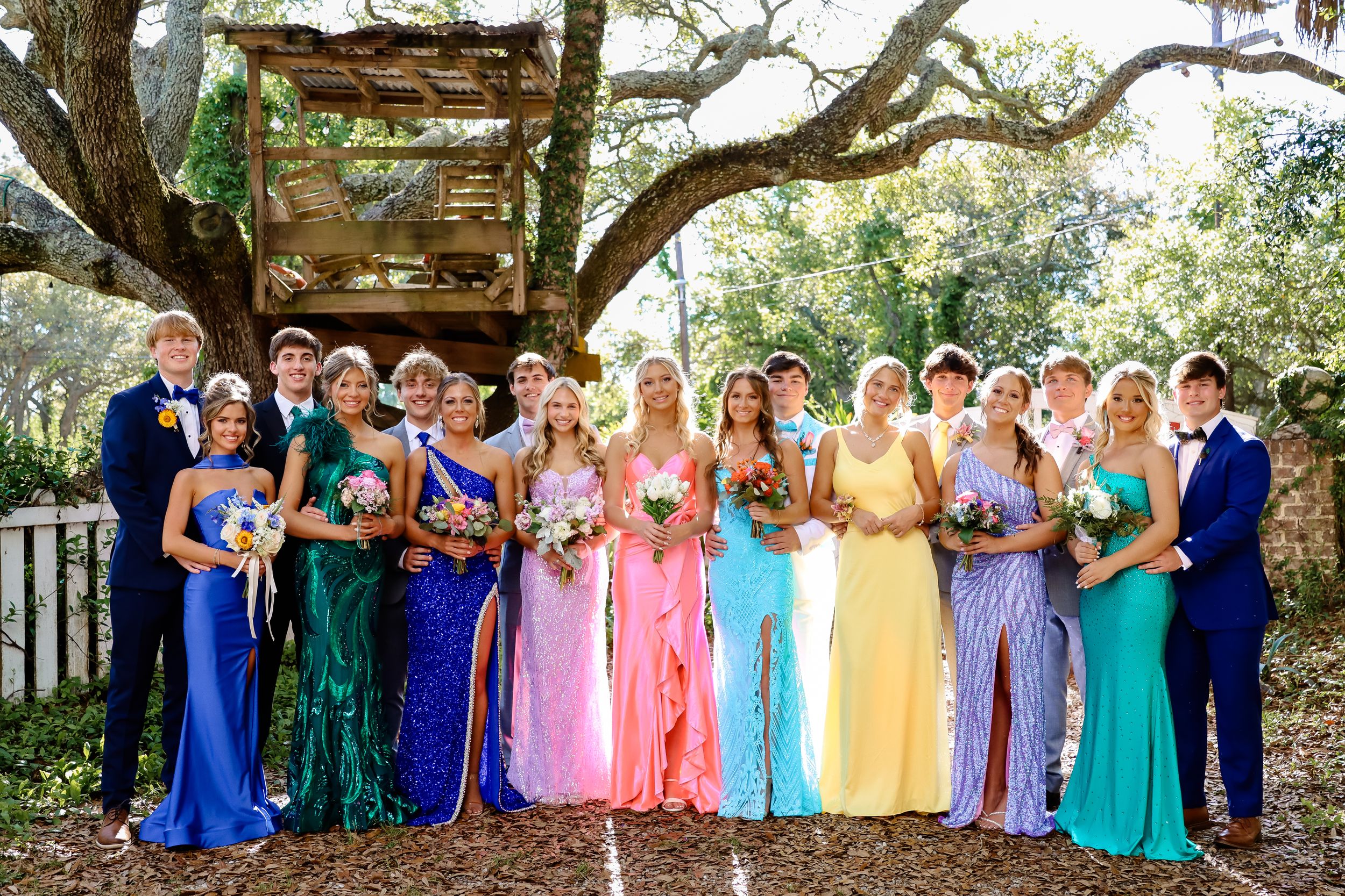 Prom party poses under oak tree wearing vibrant rainbow-colored formal dresses.