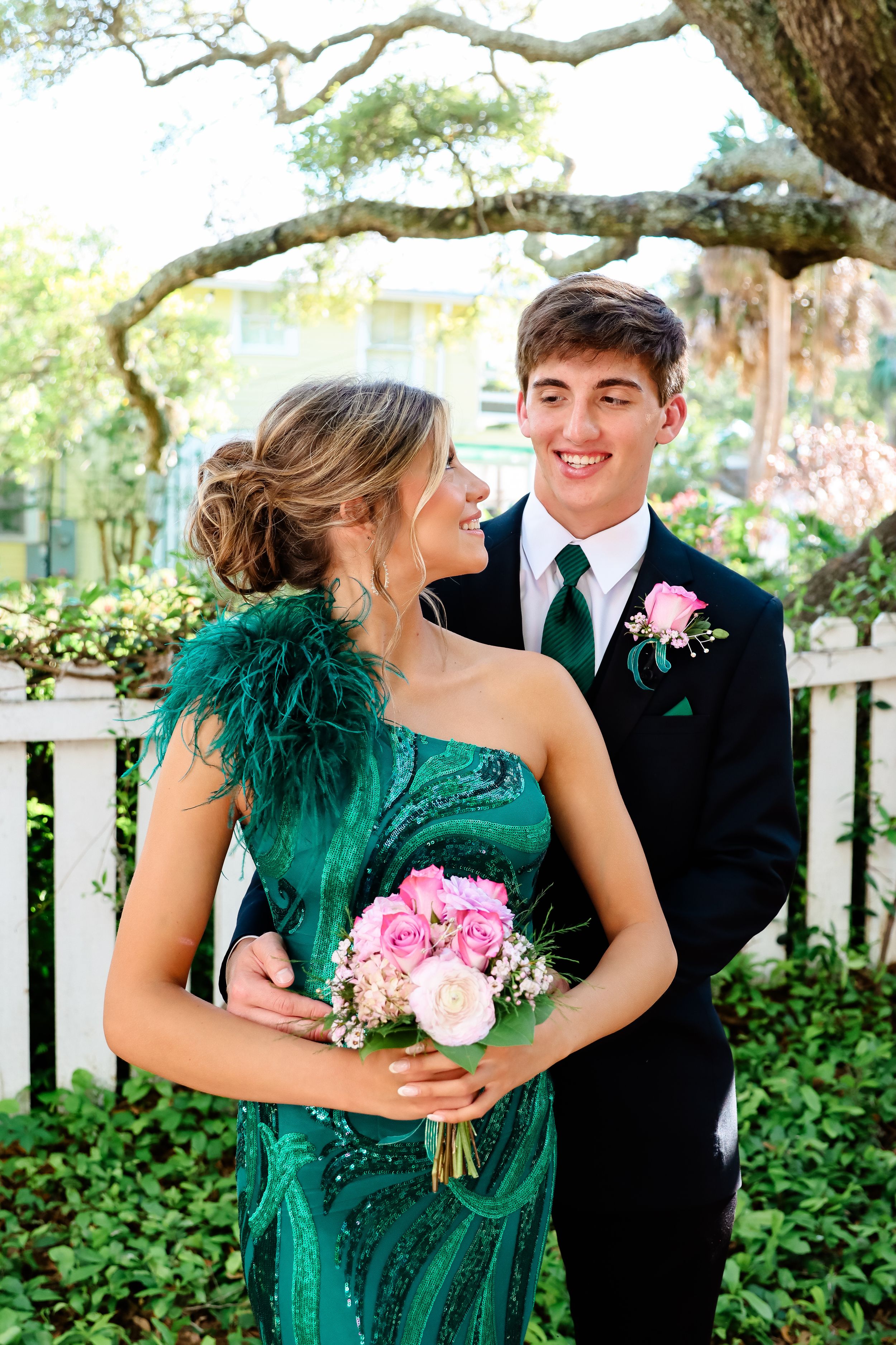 A couple poses in a garden setting, wearing formal attire of emerald green sequin dress and black tuxedo for prom or event.