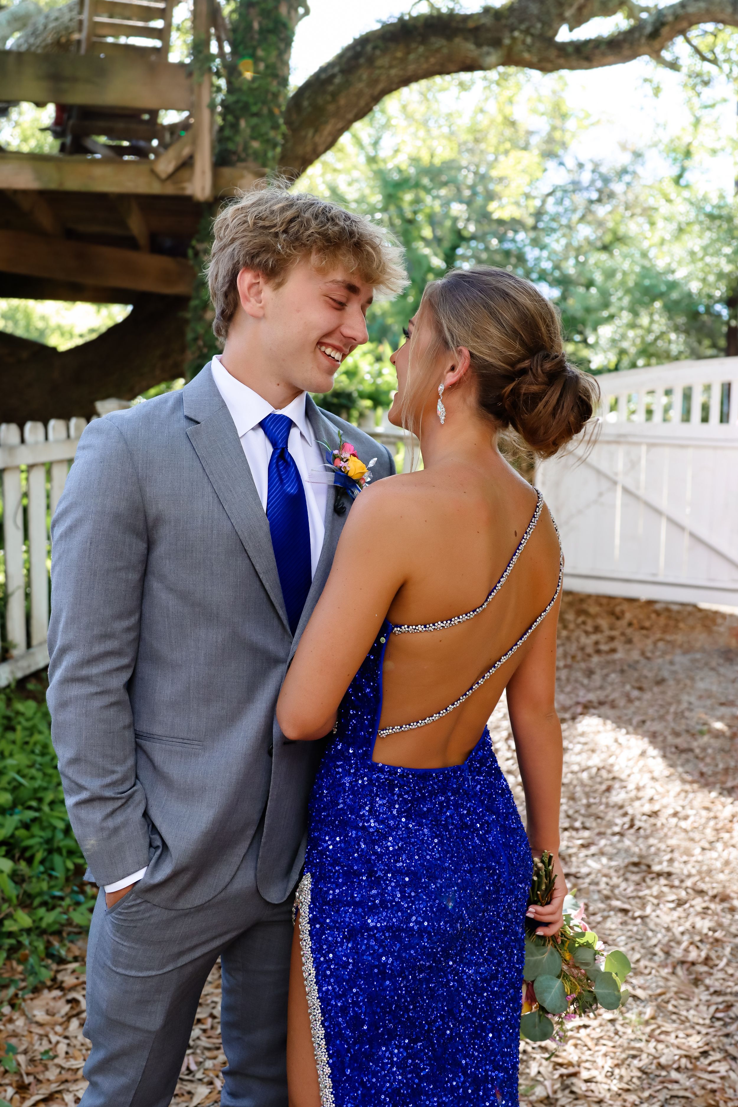 Couple in formalwear under tree canopy with person wearing sparkly royal blue backless dress and grey suit with blue tie.