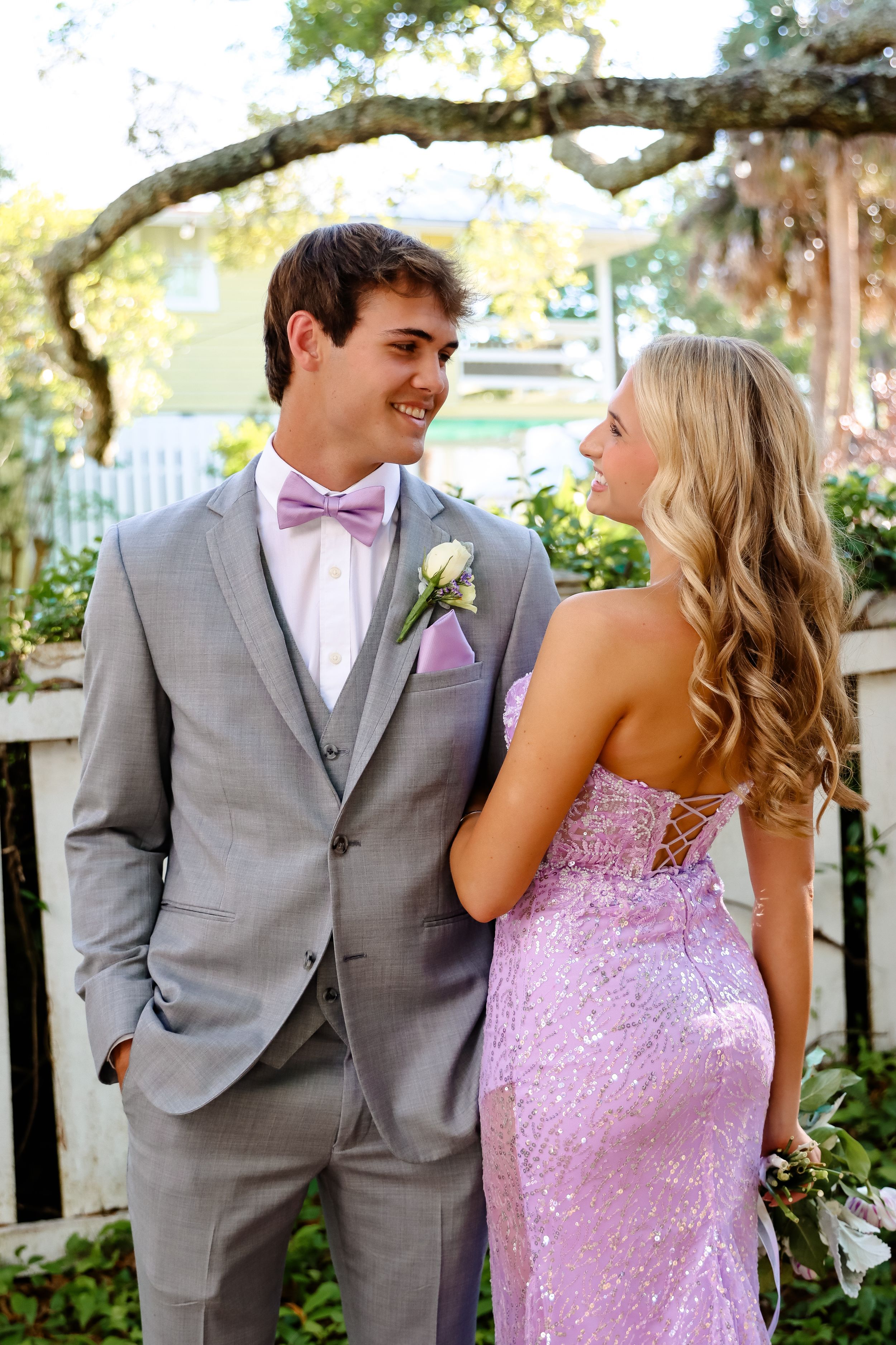 A couple in formal attire shares a sweet moment in a garden setting, with him in gray suit and her in purple sequined gown.