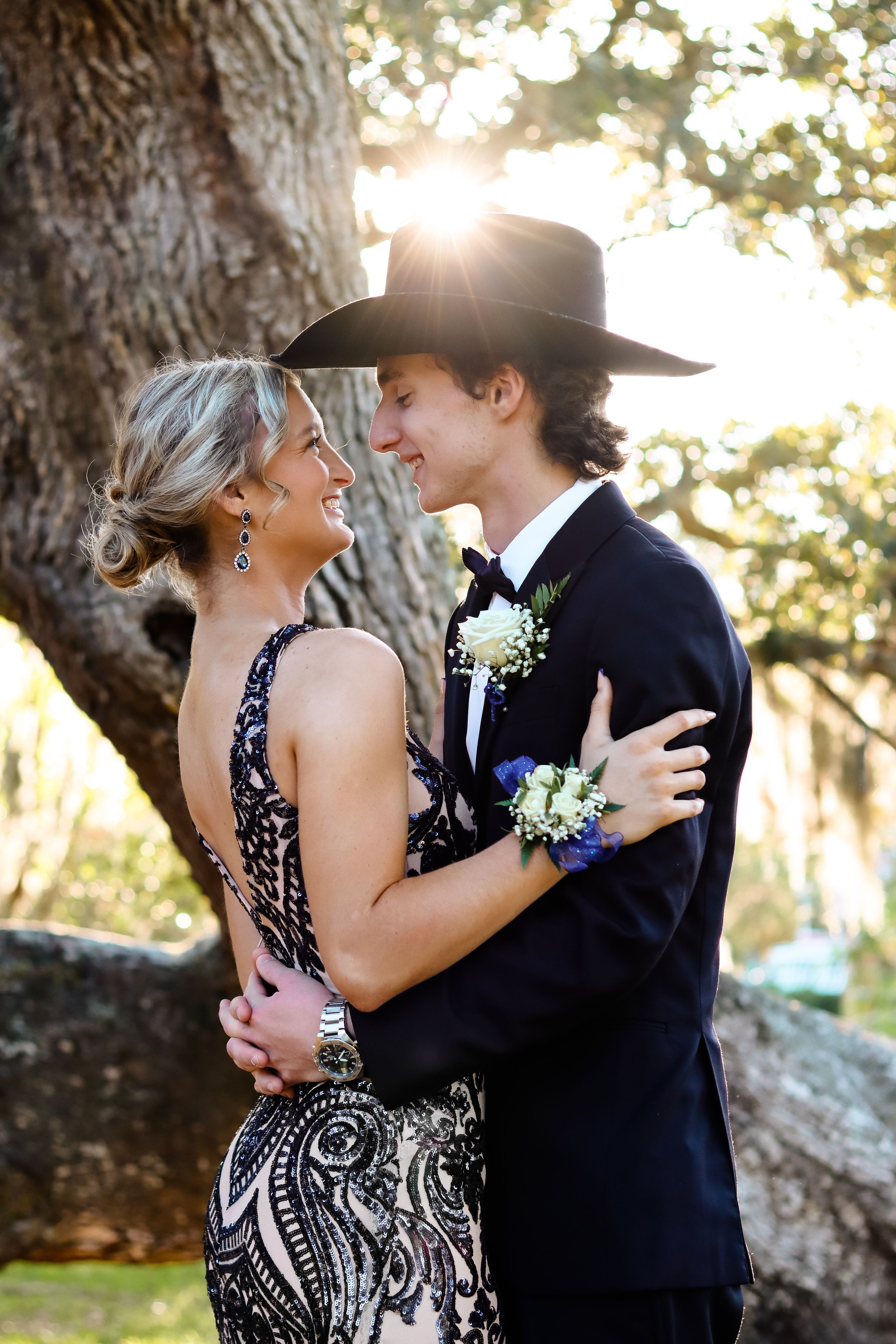 A couple shares a moment at sunset, she in a patterned dress and he in formal attire with a cowboy hat.