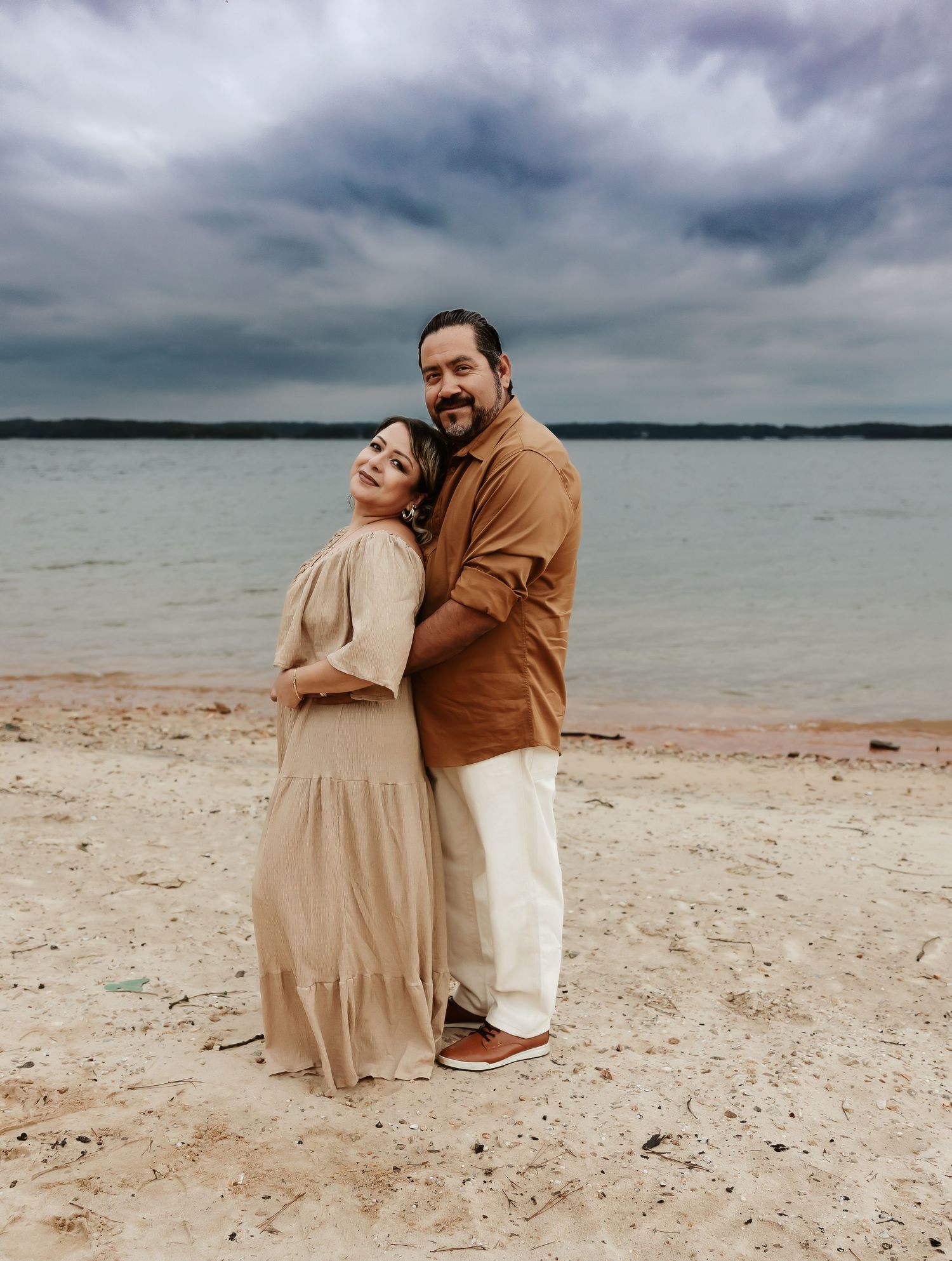 A couple in neutral tones embraces on a cloudy beach, standing against a tranquil waterfront backdrop.