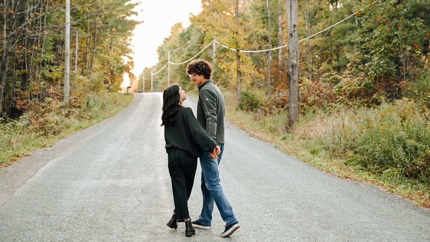 Two people walk together down a quiet rural road surrounded by autumn trees on a peaceful afternoon.