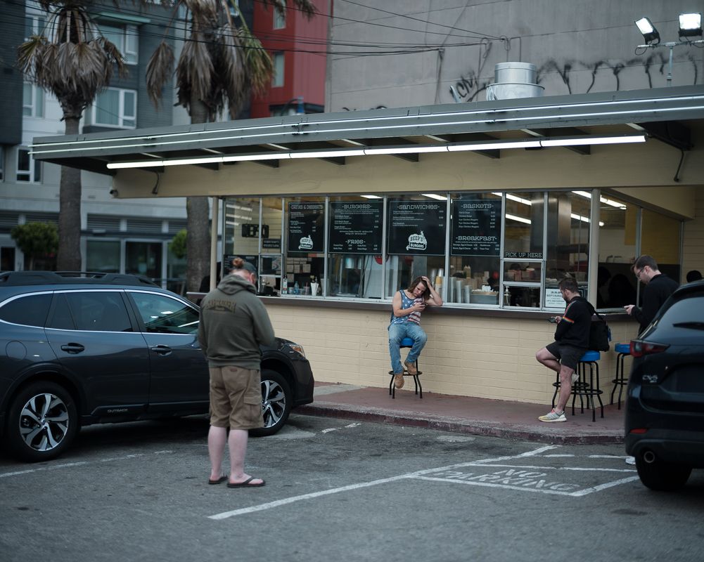 People gather at an illuminated storefront in evening with parked cars.