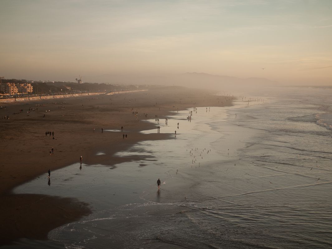 Aerial view of a misty beach at sunset with silhouettes of people scattered along the shoreline.