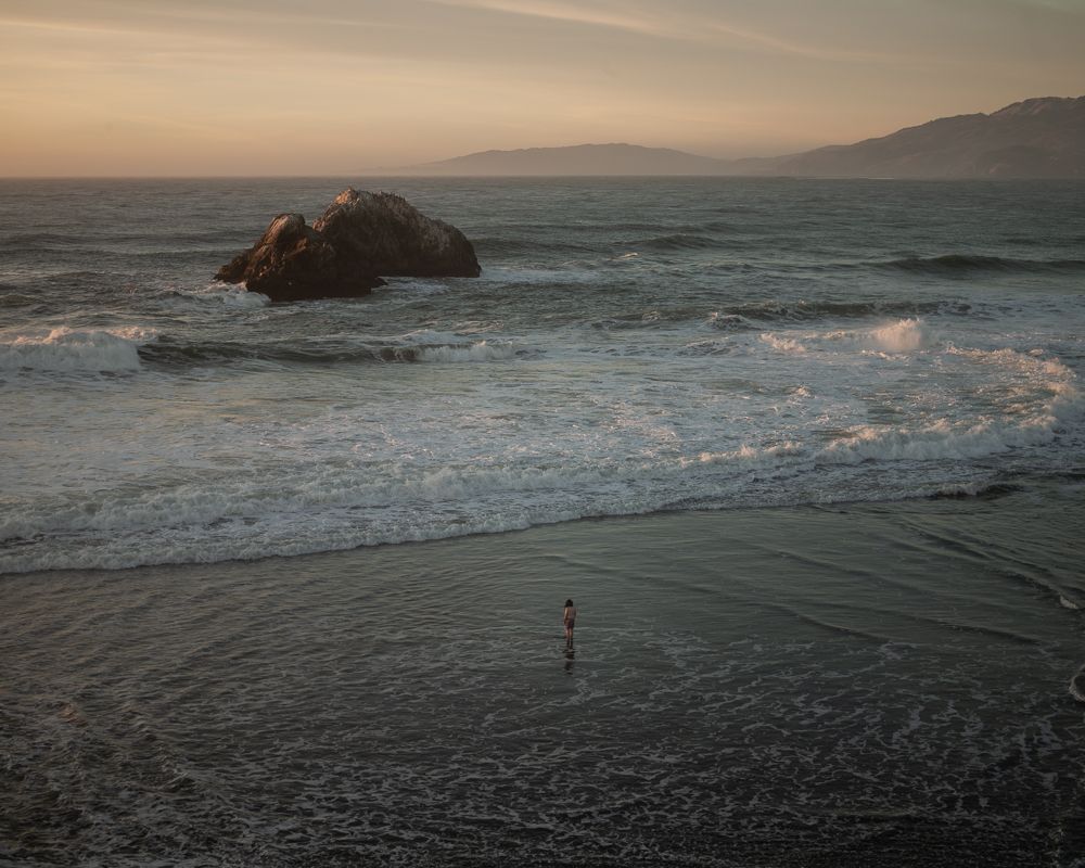Rocky outcrop in churning ocean waves at dusk with a lone figure on the beach.