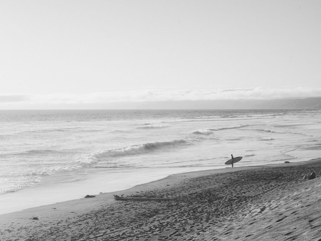 A lone surfer walks along a misty black and white beach with surfboard under cloudy skies.