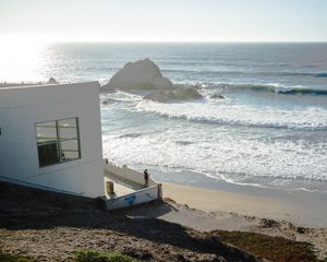 White building perched on cliff overlooking rocky coastline with crashing waves.