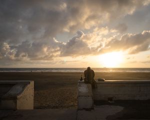 Silhouetted figure watches dramatic sunset over beach from concrete wall.