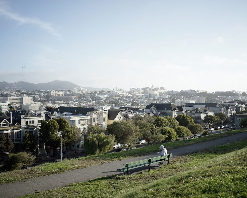 A scenic view of San Francisco cityscape from a hilltop park with someone relaxing on a bench on a sunny day.
