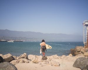 Surfer carrying board stands on rocky outcrop overlooking blue ocean bay.