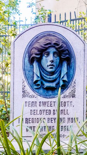 A vintage cemetery headstone with a dramatic sculptural relief medallion and ornate text against an iron fence backdrop.