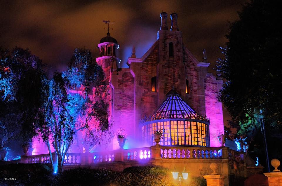 Illuminated silhouettes of haunted house buildings against a dark sky with purple and blue lighting create an eerie atmosphere.