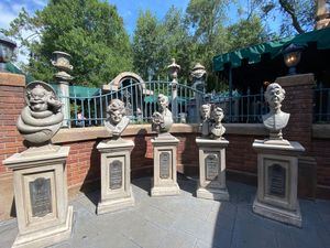 A row of decorative stone busts and sculptures line a brick wall in an outdoor garden setting.