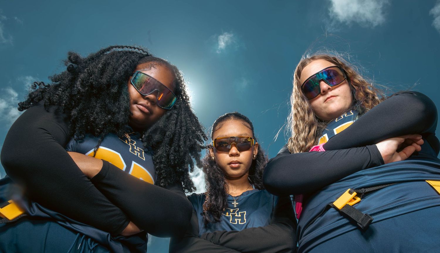 Three female athletes in blue uniforms pose confidently under a bright sky.