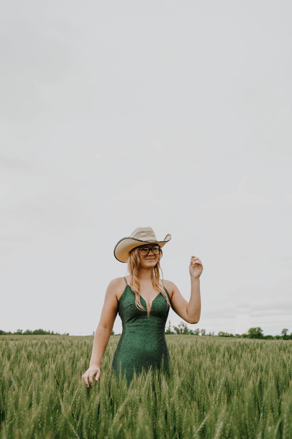 Person in a green dress and cowboy hat stands in a lush wheat field, surrounded by tall green stalks.