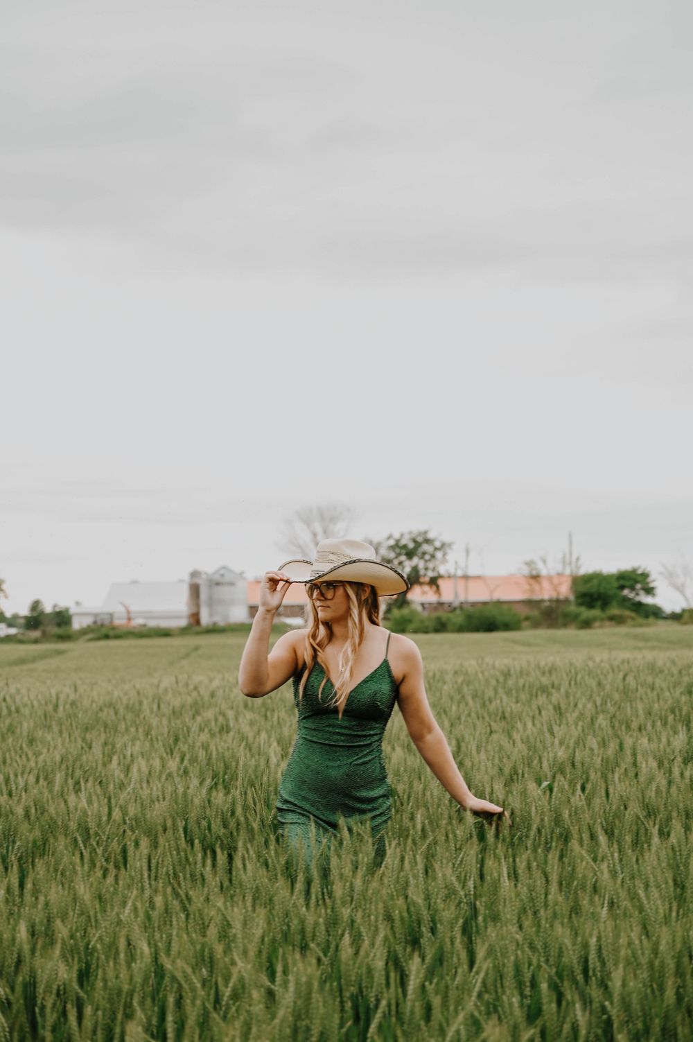 Woman in emerald dress stands among tall wheat stalks while adjusting her cowboy hat.