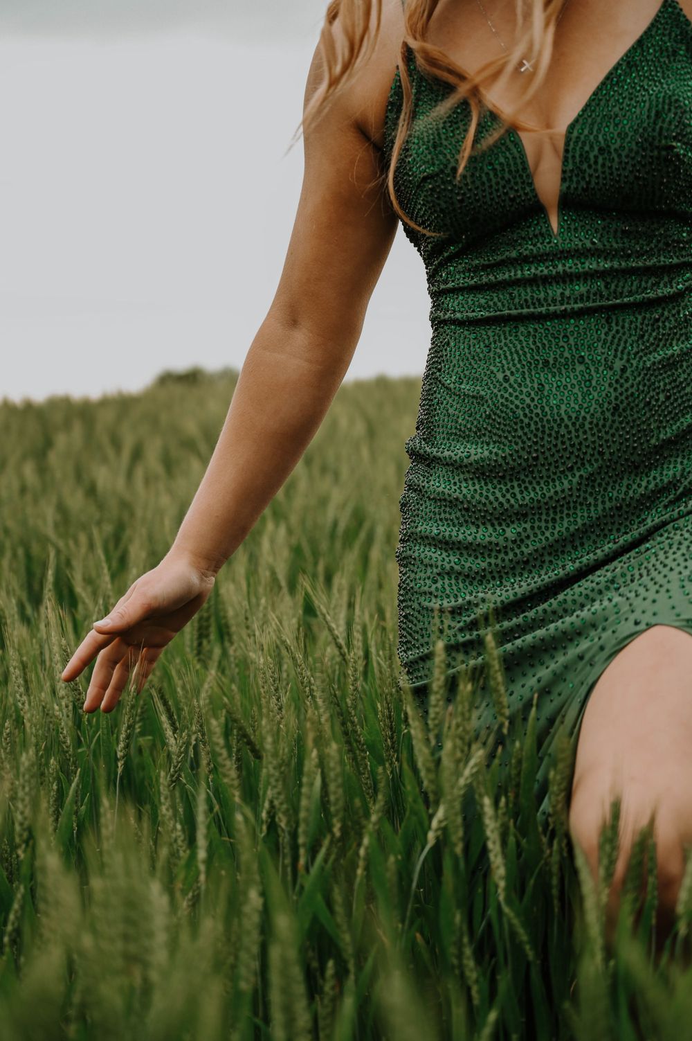 Person wearing a sparkly emerald green dress runs their hand through tall wheat stalks in a summer field.