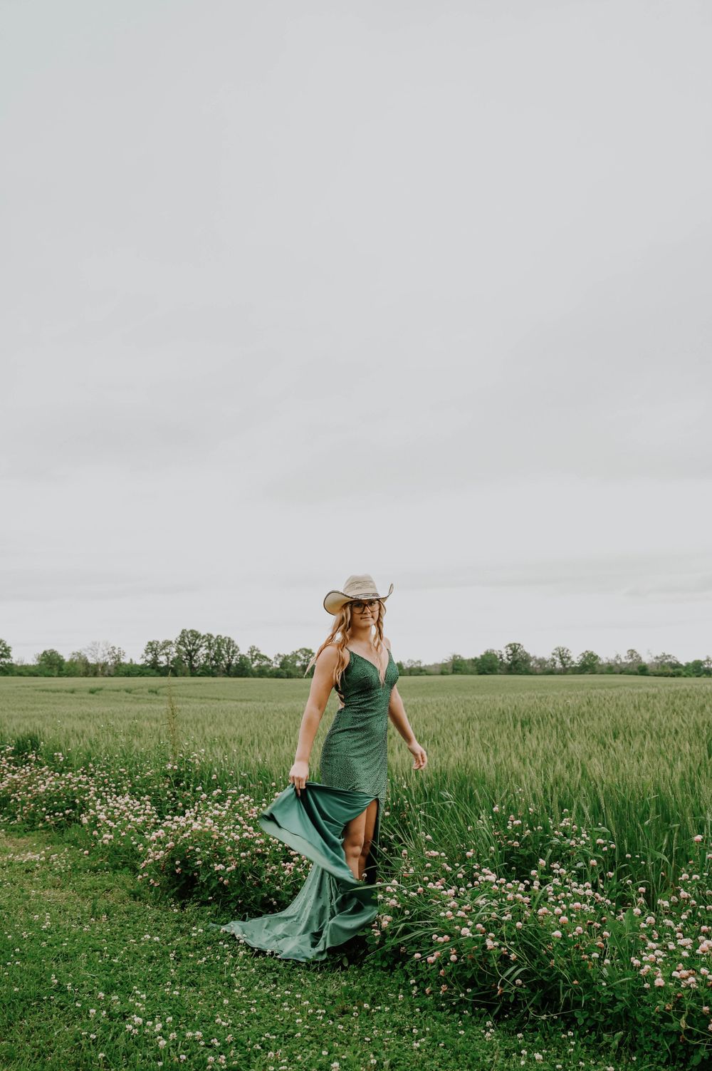 Woman in emerald dress and sun hat strolls along flowering meadow path under cloudy sky.