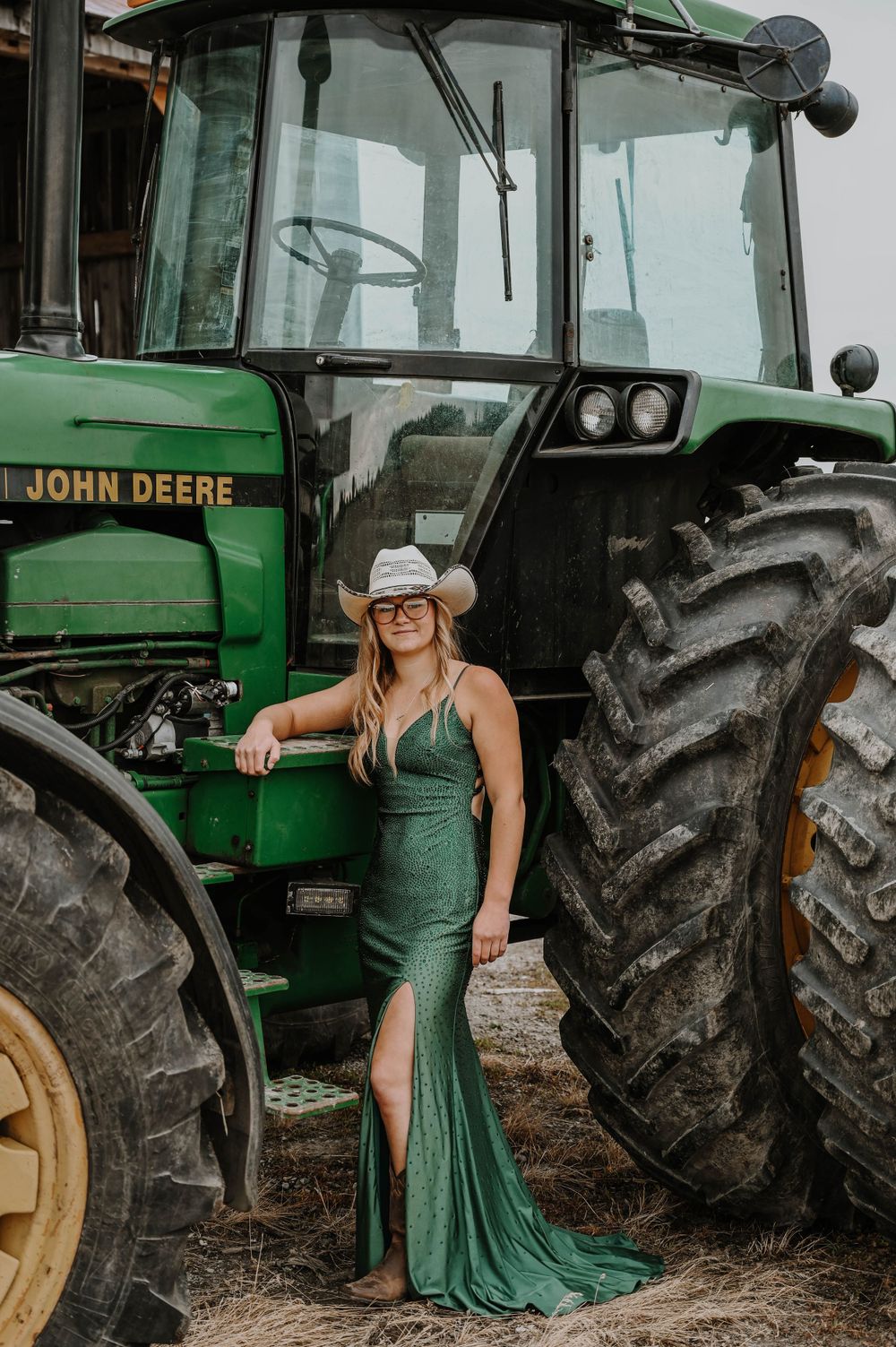 Elegant green evening gown contrasts with John Deere tractor in rustic farm photoshoot setting.