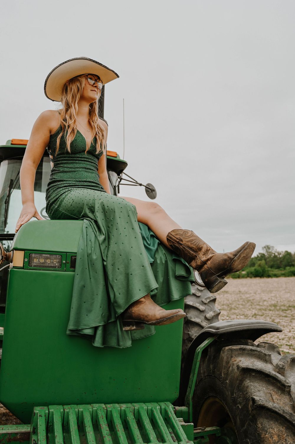 A figure in a green dress and cowboy boots sits atop a green John Deere tractor in a rustic country setting.
