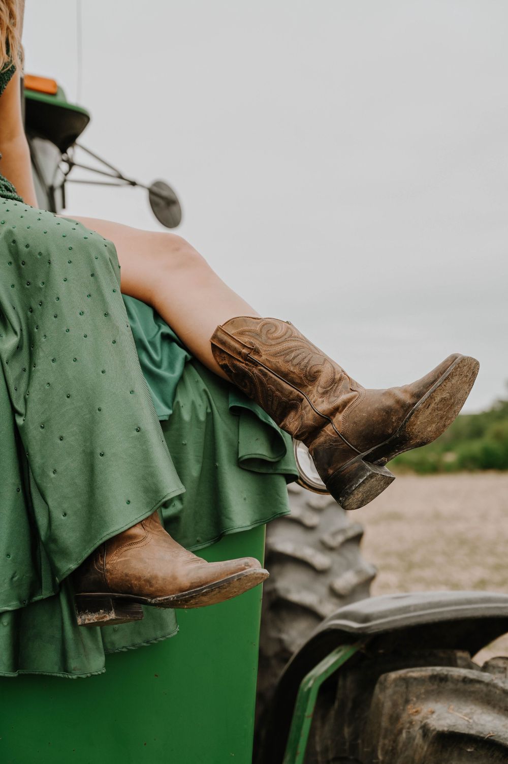 Brown cowboy boots and green polka dot dress shown against tractor tire in rustic country setting.