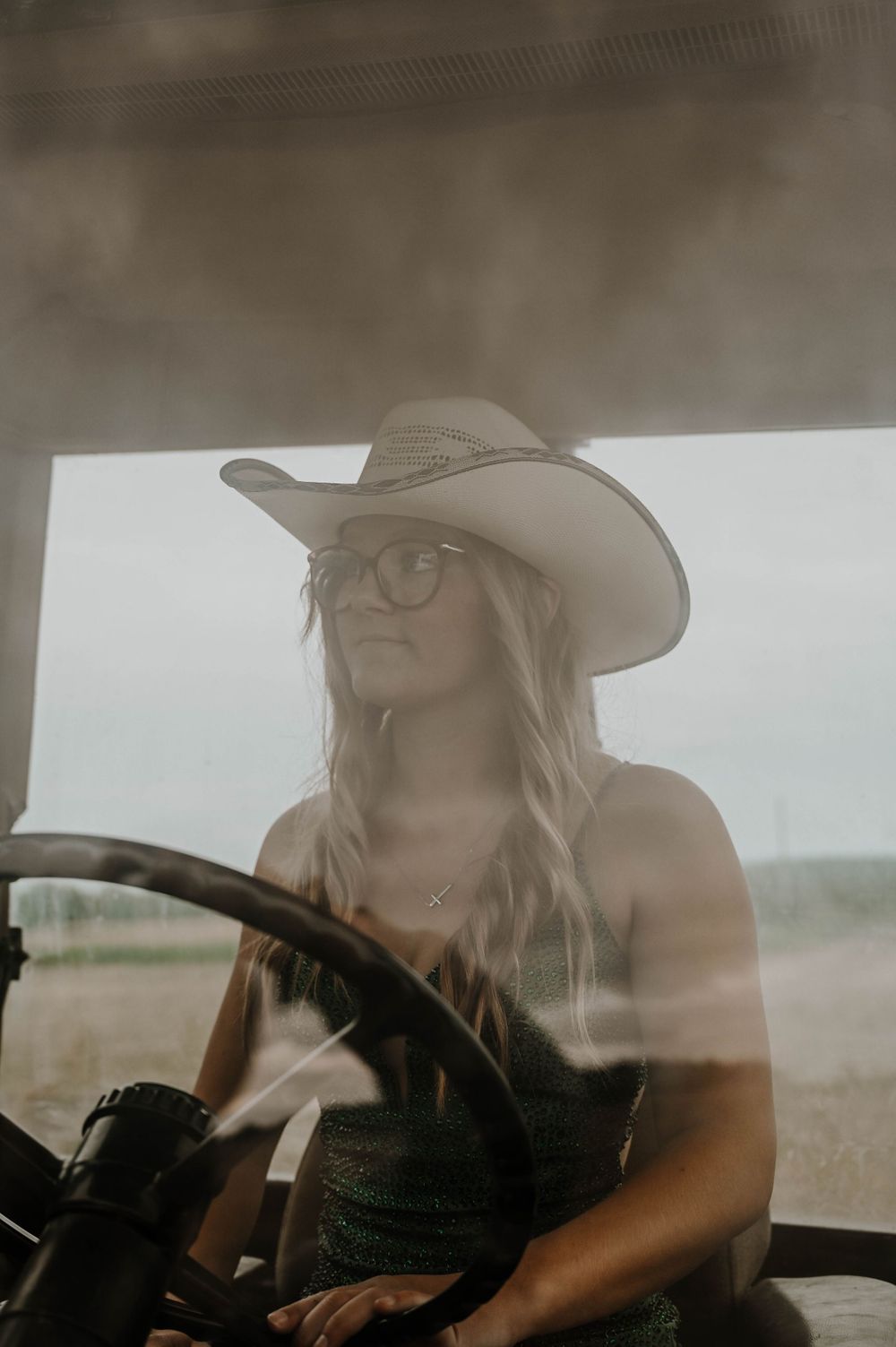 Moody sepia-toned photo shows a person in cowboy hat and glasses behind steering wheel at golden hour.