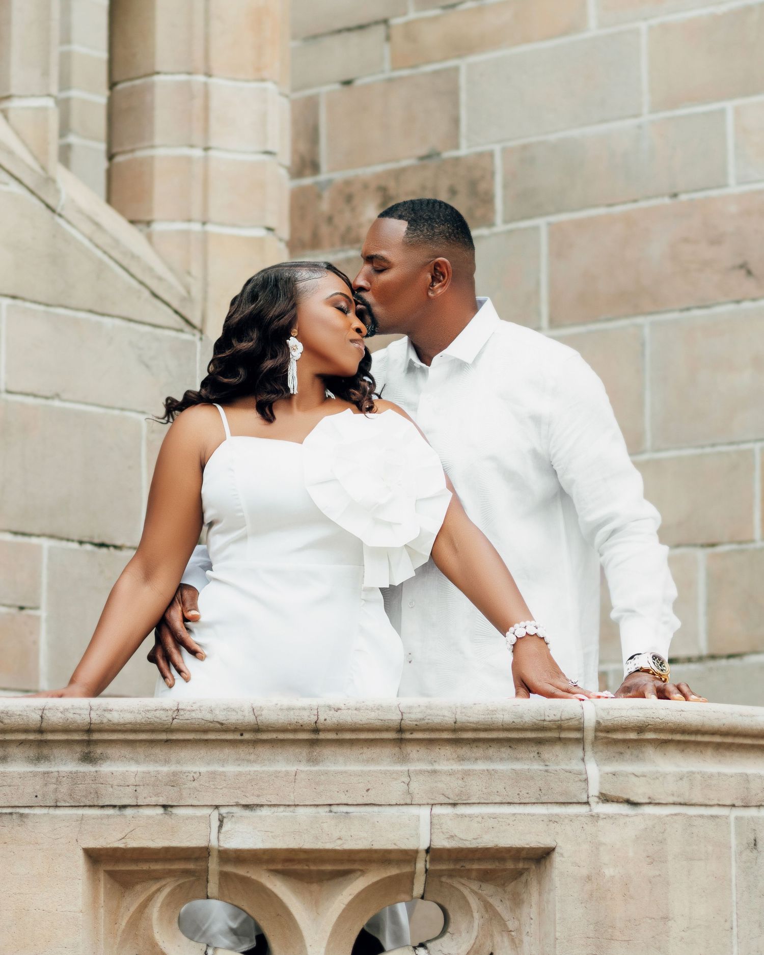 A romantic couple in matching white outfits share an intimate moment on stone steps against a vintage architectural backdrop.