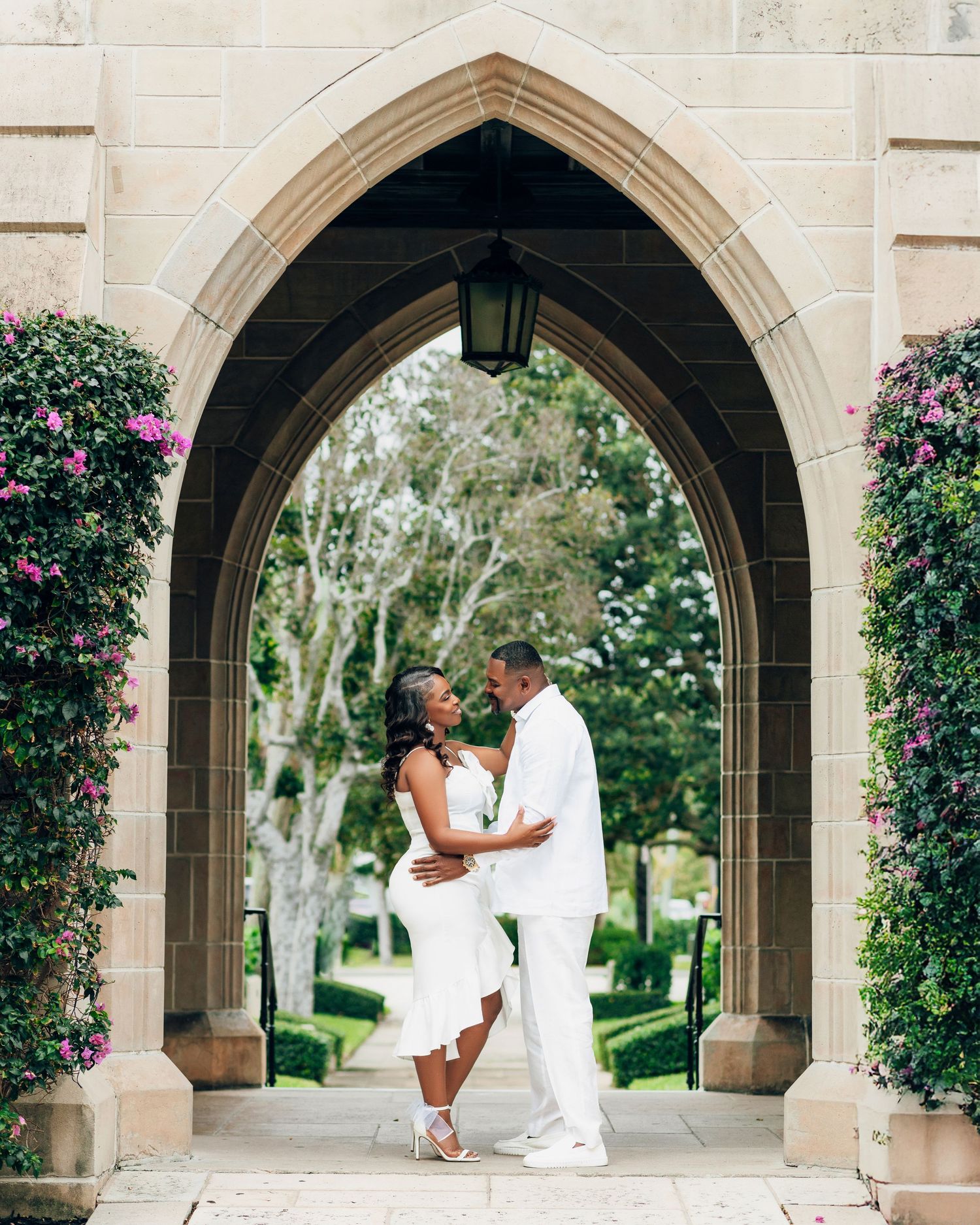 A couple embraces under a beautiful stone Gothic archway flanked by pink flowering vines in an elegant garden setting.
