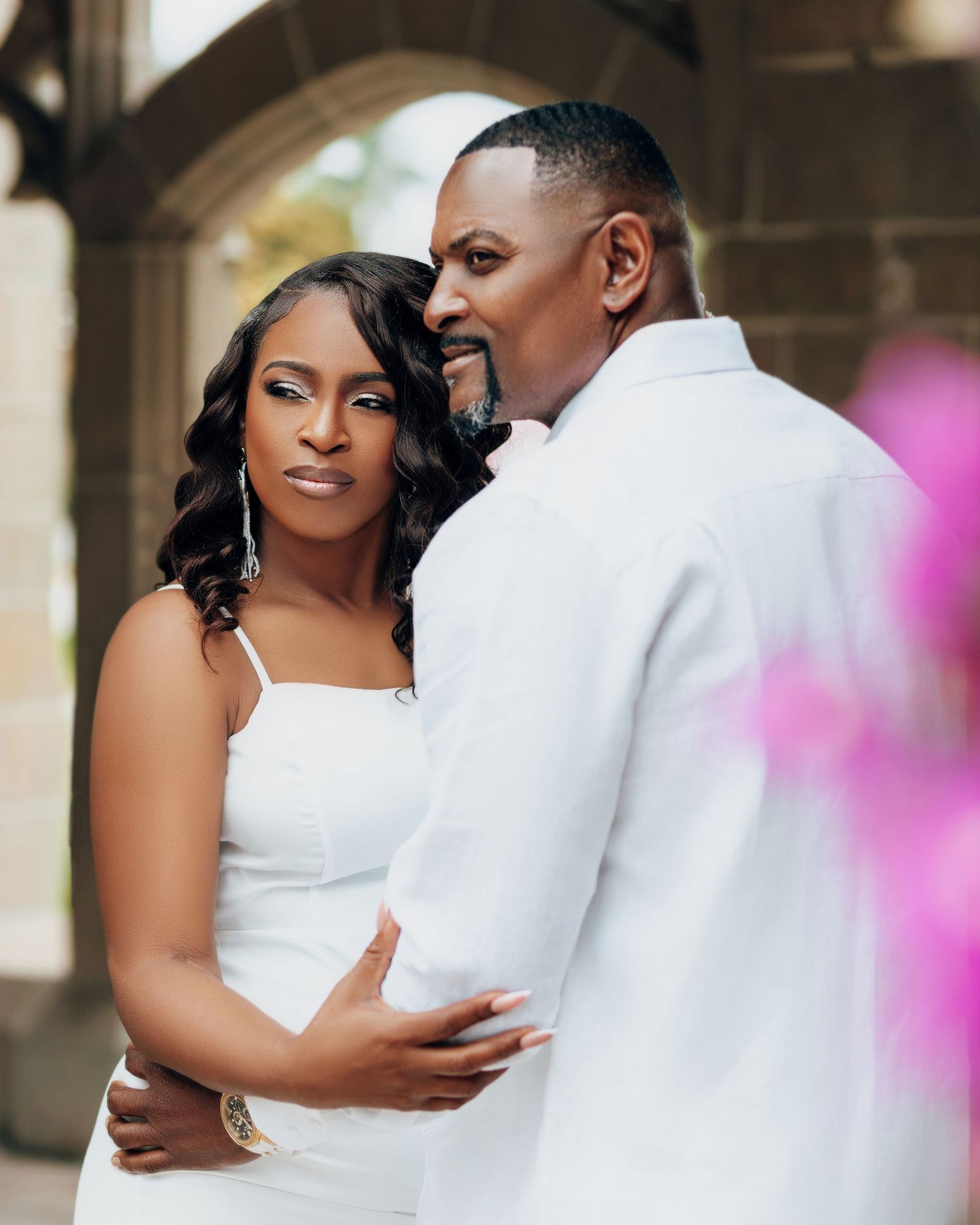 A romantic wedding portrait shows a couple in an elegant embrace with a soft pink bokeh effect in the background.