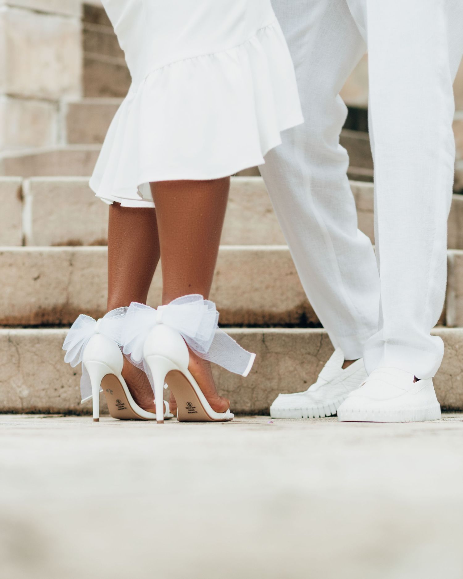 Contrasting wedding shoes shown between couple standing on steps - elegant white bridal heels and casual white sneakers.