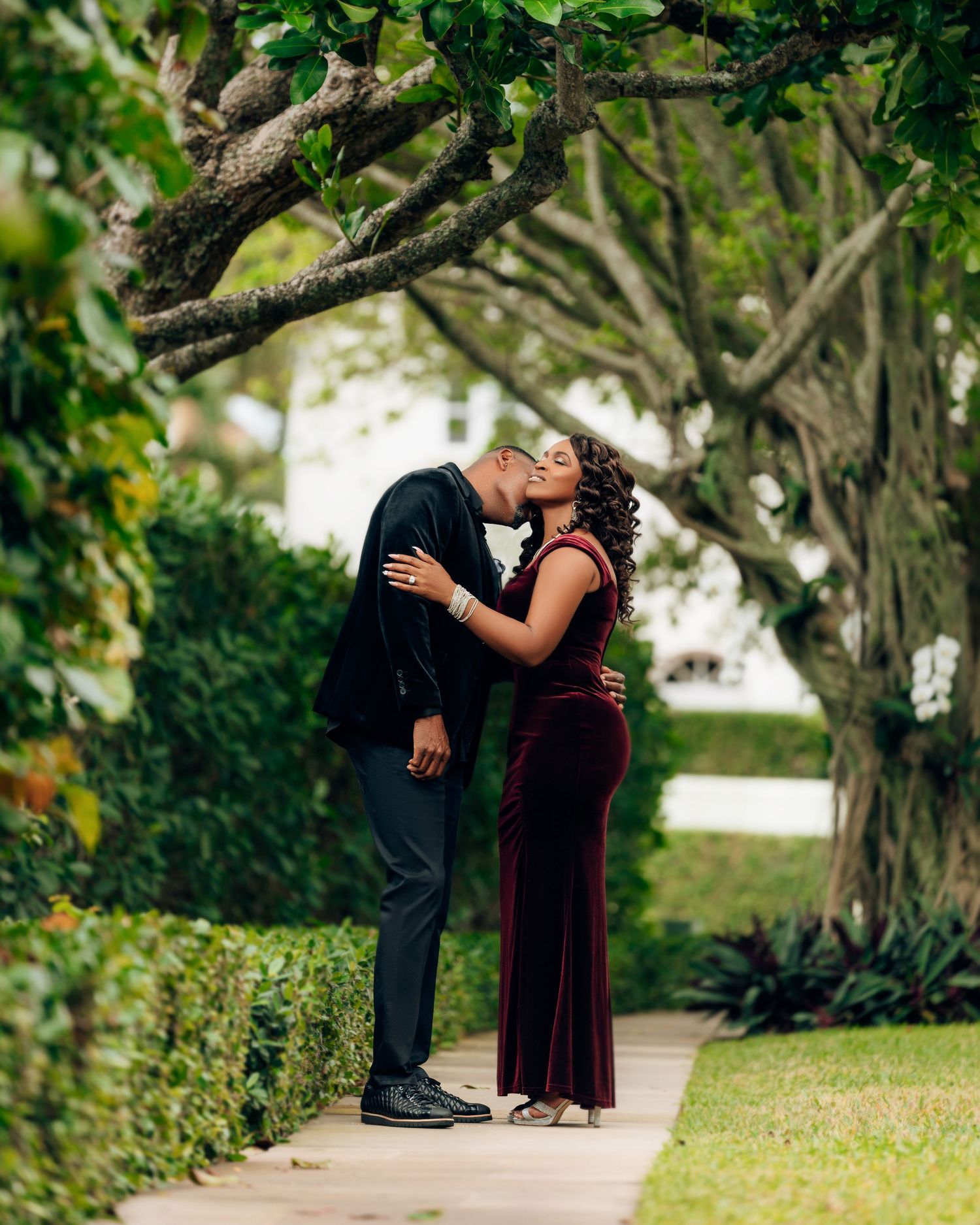 A romantic couple embraces on a garden path surrounded by lush trees and greenery in an elegant outdoor setting.