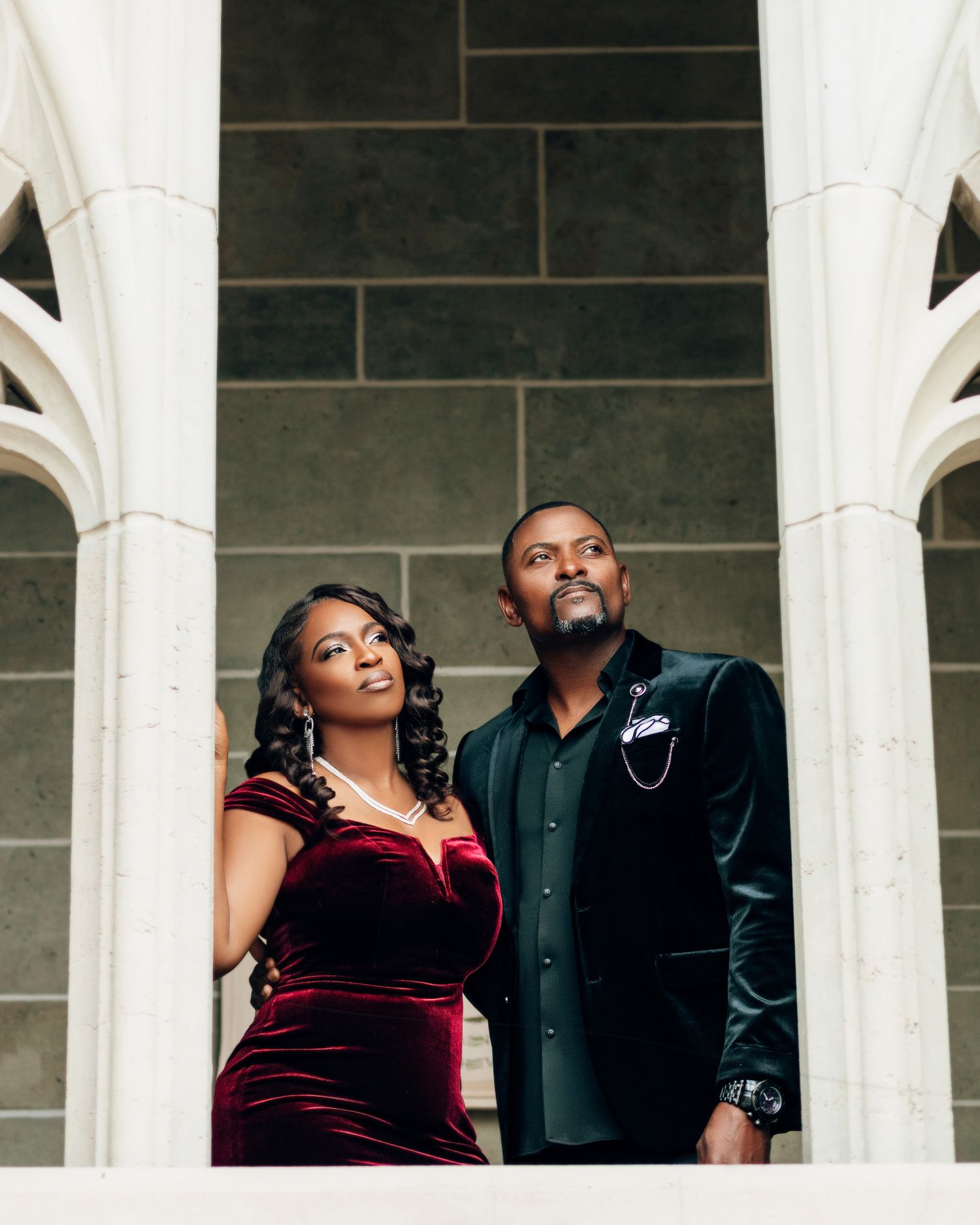 A couple in formal attire poses between white stone columns, wearing burgundy velvet dress and navy velvet blazer.