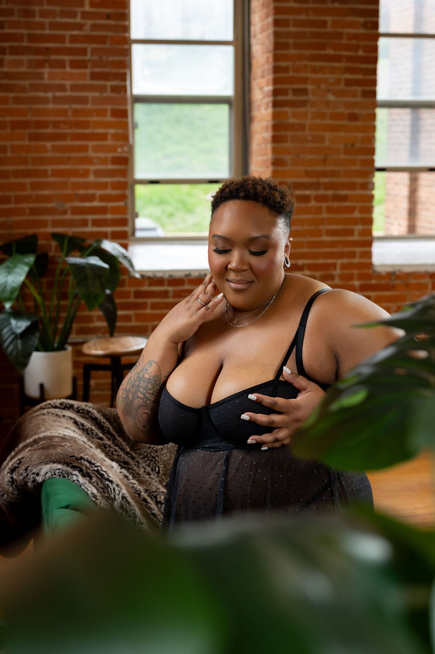 Plus-size woman in black bra sitting in chair near brick wall surrounded by plants.