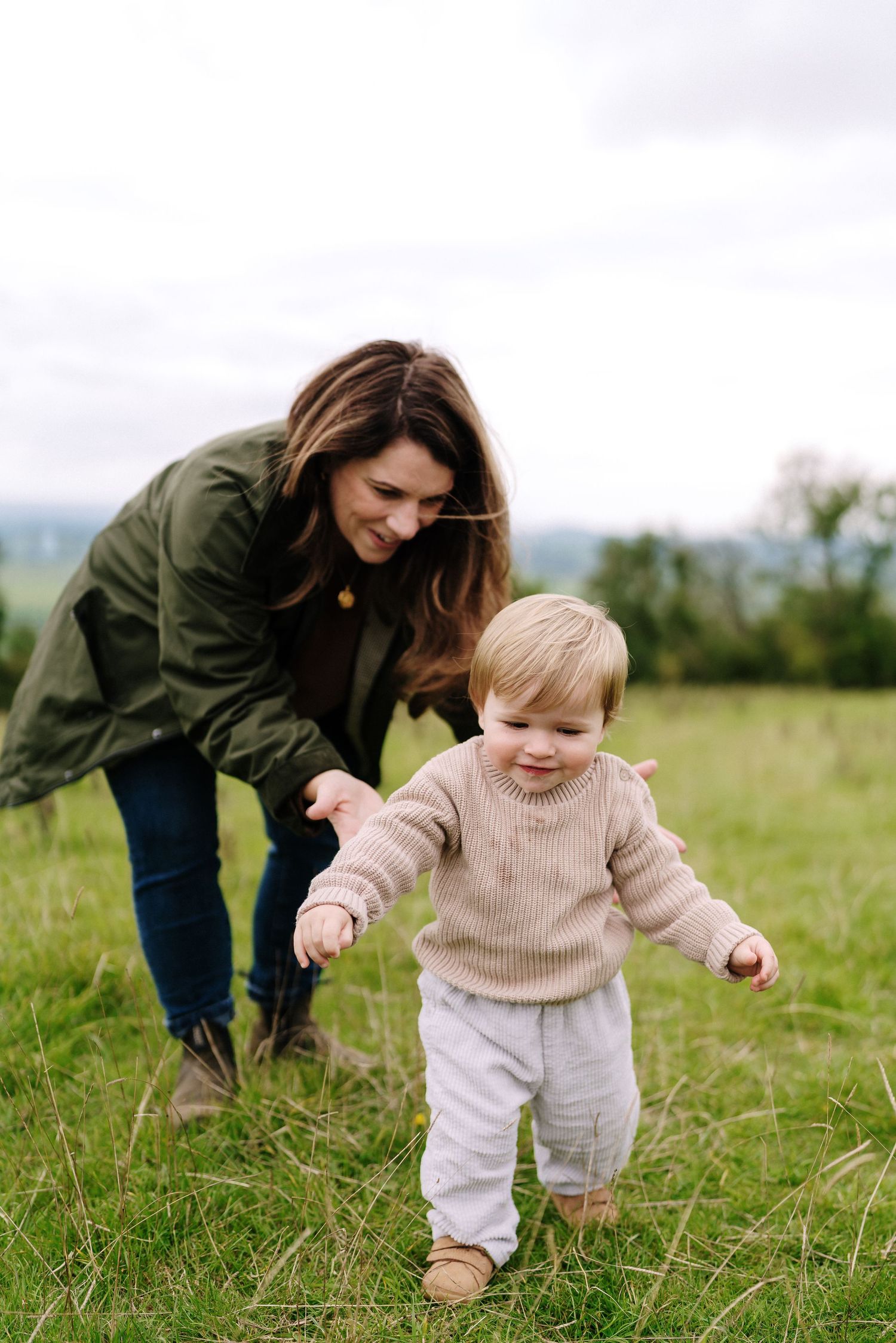 A toddler in a beige sweater takes first steps across a grassy field while being supported from behind.